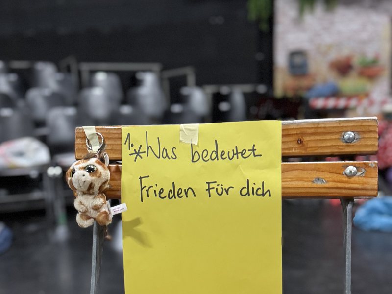 Ein gelber Zettel auf einer Stuhllehne mit der handgeschriebenen Frage: Was bedeutet Frieden für dich? Daneben ein kleines Stofftier